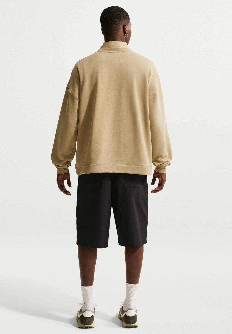 Uomo in piedi di spalle, indossa una maglietta beige a maniche lunghe, pantaloncini neri, calzini bianchi e sneakers bianche su sfondo chiaro uniforme.