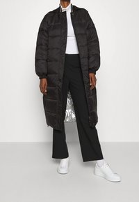 Lang sort pufferjakke med hætte og ribbet krave, med lynlåse foran og sølvfor. Båret sammen med sorte bukser og hvide sneakers.