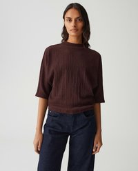 Frau in einem strukturierten, dunkelbraunen Kurzarmoberteil und dunkelblauen Jeans, die vor einem einfarbigen, hellen Hintergrund steht.
