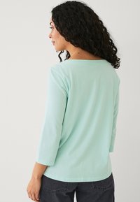 Maglietta a maniche lunghe di un verde menta chiaro con una ampia scollatura a barca, realizzata in morbido cotone, con una vestibilità rilassata e un sottile dettaglio di cucitura sul retro.