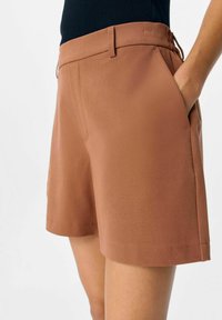 Shorts marron sur mesure en tissu lisse, avec une taille plate, des poches latérales et une coupe décontractée. La ceinture comprend un cordon de serrage réglable.