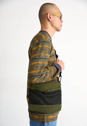 Grønn budveske med svart mesh-front, som har en glidelåslomme. Teksturlaget og justerbar skulderrem er synlig.