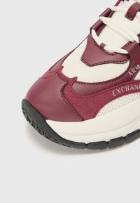 Baskets de sport bordeaux et crème avec un dessus en maille texturée, une semelle en caoutchouc et des éléments de design contrastés avec une marque bien en vue.