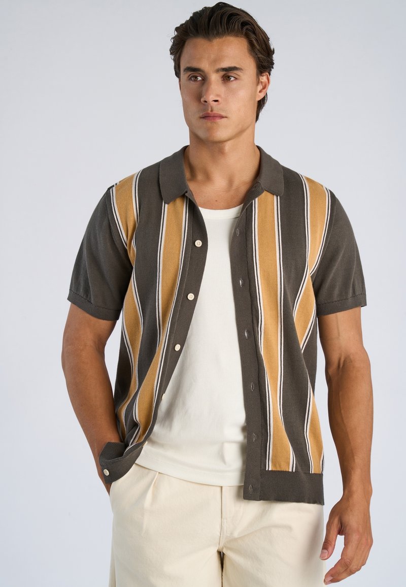 Giovane uomo con pantaloni beige, canottiera bianca a coste e camicia a maniche corte aperta con righe verticali marroni e gialle.