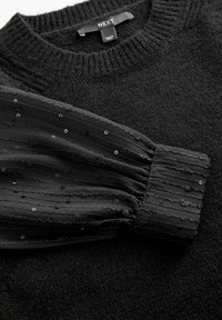 Pull noir comprenant un corps texturé en tricot et une manche plissée transparente agrémentée de sequins. L'étiquette porte le nom de la marque.