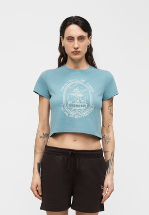 Femme aux cheveux foncés et tatouages, portant un T-shirt bleu court avec un motif de fontaine et un short noir à cordon, se tenant devant un fond blanc.
