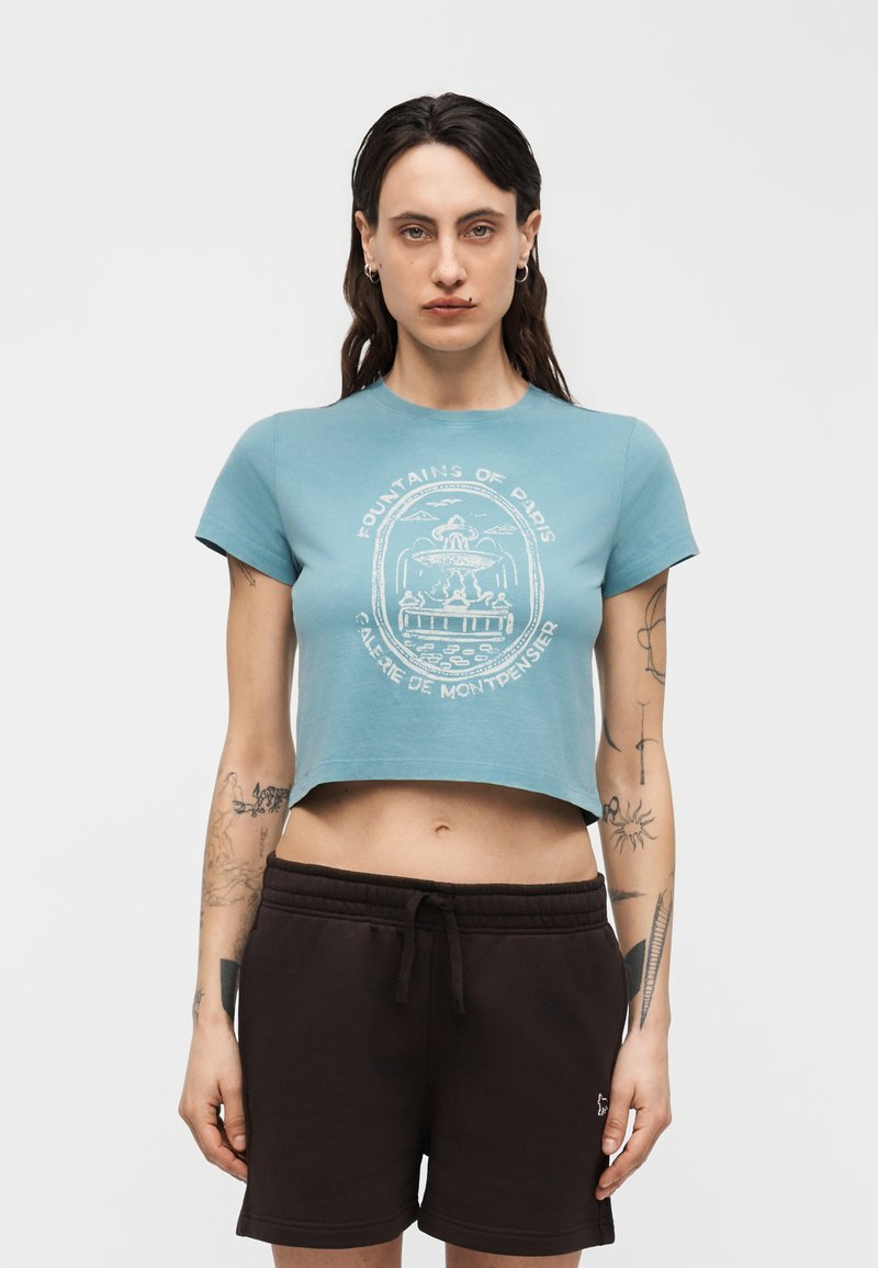 Γυναίκα με σκούρα μαλλιά και τατουάζ, ντυμένη με μπλε κοντομάνικο T-shirt με γραφικό συντριβάνι και μαύρο σορτς με κορδόνι, στέκεται μπροστά σε λευκό φόντο.