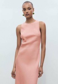 Abito senza maniche, aderente, in morbido tessuto rosa con una consistenza liscia. La modella accessoriza con grandi orecchini verdi. Design semplice ed elegante.