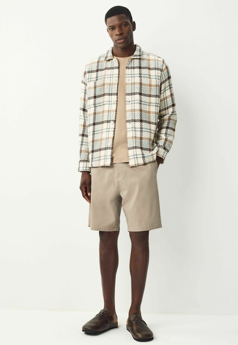Homme portant un short beige, une chemise beige, une veste à carreaux boutonnée et des chaussures noires à enfiler, se tenant devant un fond blanc uni.