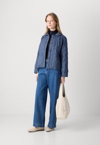Marineblauwe gewatteerde jas met knoopsluiting, gecombineerd met lichtblauwe wijde jeans, beige flats en een slonterige handtas in neutrale tinten.