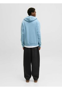 Sudadera azul claro con un corte relajado y bolsillo frontal, combinada con pantalones negros de corte holgado y zapatos marrones. Textura plana, diseño minimalista.