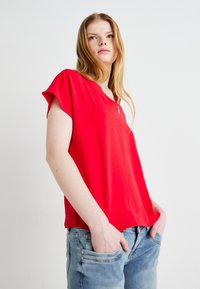 Red short-sleeve blouse with a V-neckline, made from a smooth fabric. Worn with light blue denim jeans, hands in pockets. Simple design.