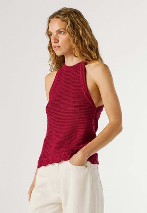 Žena s valovitom plavom kosom, odjevena u crveni pleteni top bez rukava s visokim okovratnikom i bijele hlače, stoji s jednom rukom u džepu, gleda lijevo.