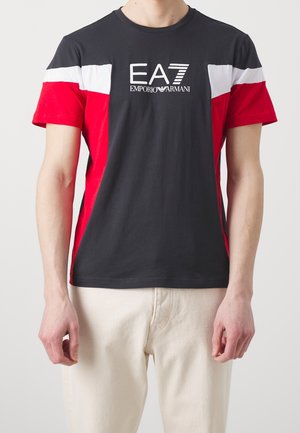 Uomo che indossa una maglietta a maniche corte nera, rossa e bianca con il logo "EA7 Emporio Armani" sul petto e pantaloni chiari.