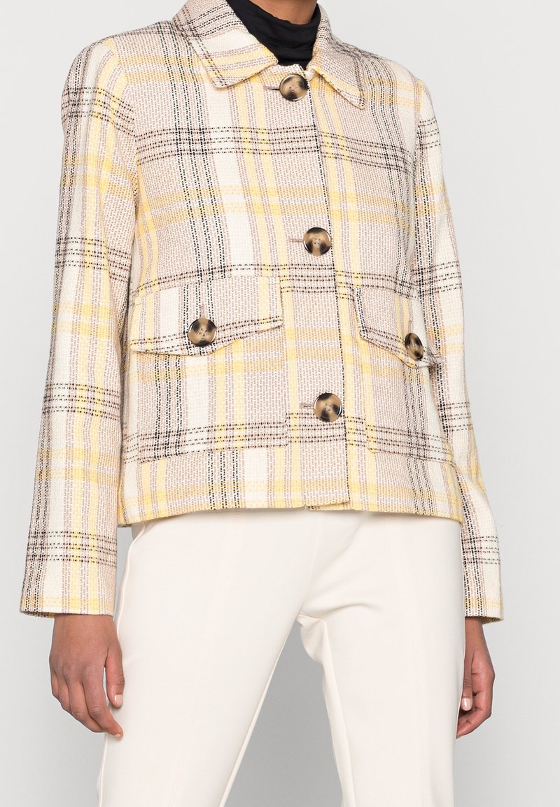 Femme portant une veste boutonnée à carreaux beige, jaune et noir avec deux poches avant, associée à un pantalon blanc.
