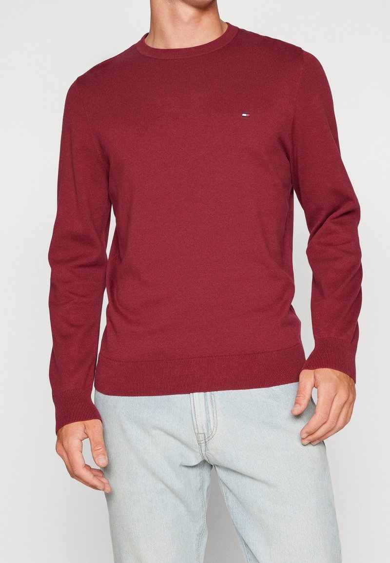 Pull en maille rouge à manches longues, encolure ronde, poignets et ourlet côtelés, avec un petit logo sur la poitrine. Porté avec un jean bleu clair.