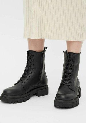 Botas de combate de cuero negro con diseño de cordones, cierre de cremallera, suela de goma gruesa y textura suave, presentadas con una falda beige acanalada.