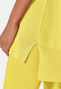 Žlutý pletený top s rozparkem na lemu a žebrovanými detaily, sladěný s žlutými kalhotami. Záběr zblízka na texturu látky a její design.