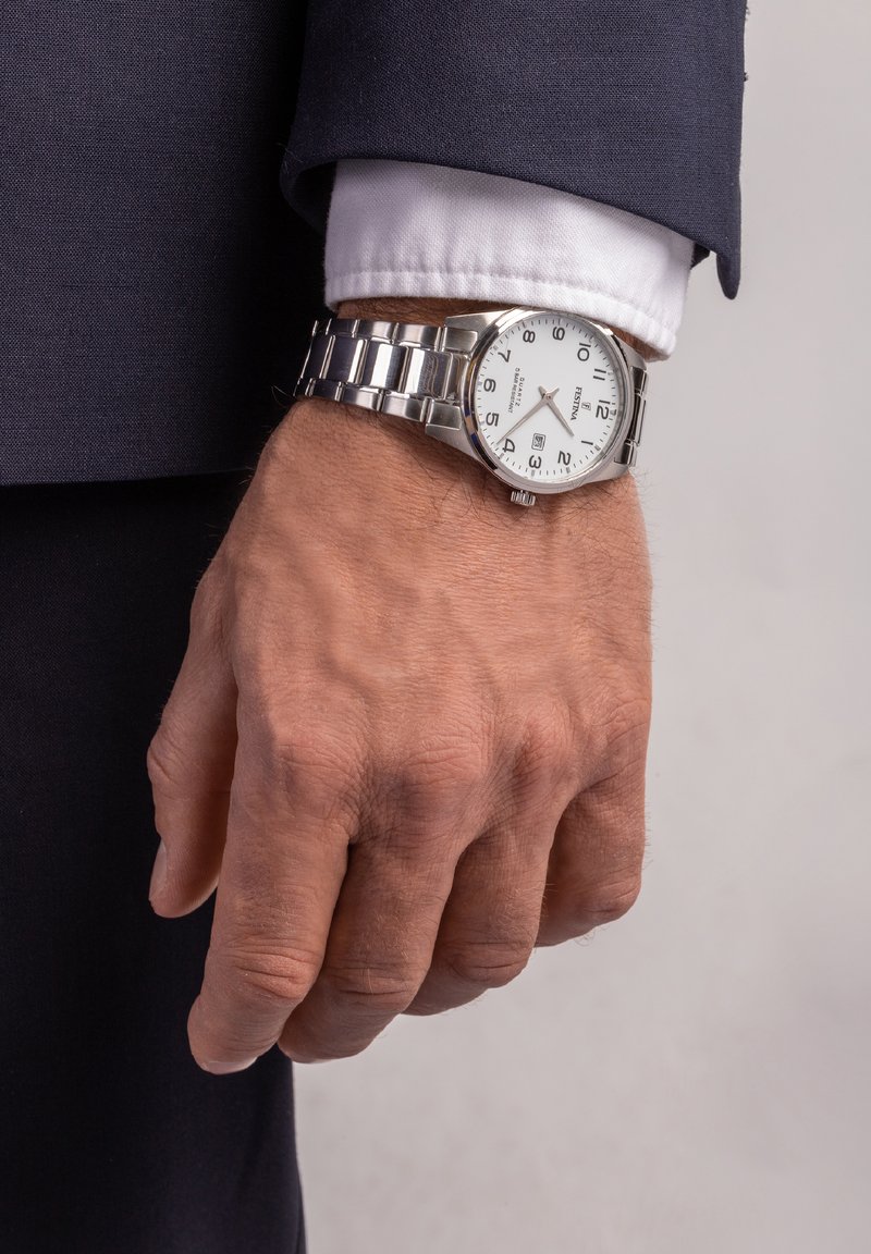 Montre en acier inoxydable avec un cadran analogique blanc, des chiffres noirs et un affichage de la date, port�ée au poignet dans un costume sombre.