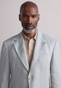 Blazer azzurro chiaro in lino con rever a lancia e chiusura a un solo bottone, indossato sopra una polo marrone chiaro a coste con tre bottoni.