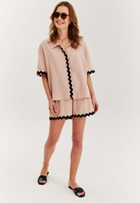 Svetloružový krátky rukáv top a zodpovedajúce šortky s čiernym ozdobným lemom. Modelka nosí čierne šľapky a slnečné okuliare.