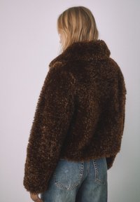 Brun jacka i konstpäls med bred krage och lös passform, kombinerad med ljusblå jeans. Texturen ser mjuk och fluffig ut.