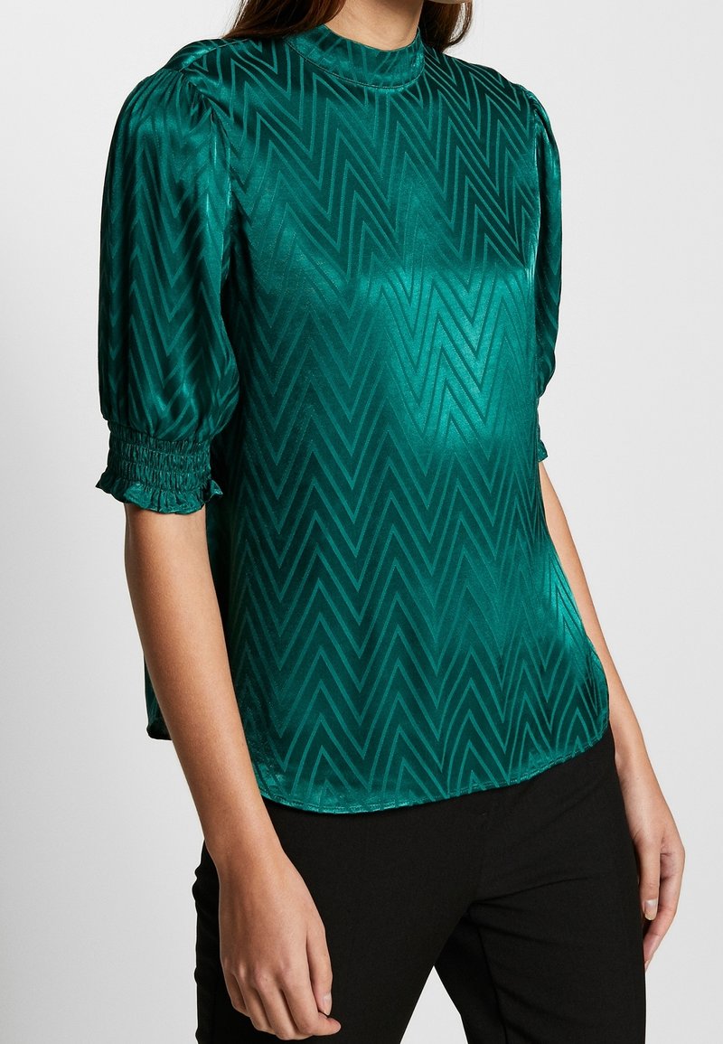 Blouse vert émeraude avec un col haut, manches courtes bouffantes et motif en zigzag. Fabriquée en satin, avec une texture lisse et brillante.
