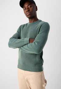 Pull en maille côtelée vert avec un col rond et des manches longues, présentant un tissu texturé et un ourlet ajusté. Porté avec un pantalon beige clair.