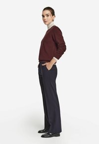 Burgunder Pullover mit langen Ärmeln und Rundhalsausschnitt, kombiniert mit marineblauen weiten Hosen. Glänzende schwarze Schuhe runden das Outfit ab.