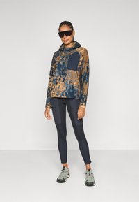 Hooded, tie-dye pullover in bruin en marineblauw, met een effen marineblauwe zak; gecombineerd met marineblauwe leggings met textuur en grijze sneakers.