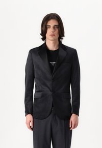 Blazer negro de tejido suave con solapas de satén, dos bolsillos frontales y cierre de un solo botón, usado sobre una camiseta negra.