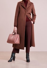 Nainen, jolla on päällään ruskea pitkä takki burgundin värisen poolopaidan ja housujen päällä, pitää pinkkiä käsilaukkua ja hänellä on mustat korkeat nilkkurit.