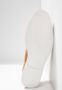 Chaussure en suede marron clair avec une semelle en caoutchouc blanc au surface texturé, inclinée contre un fond gris clair pour le contraste.