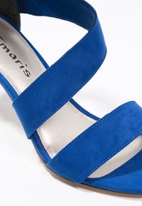 Sandalo open-toe blu scamosciato con due ampie cinghie che attraversano il piede e una soletta interna argentata su uno sfondo bianco.
