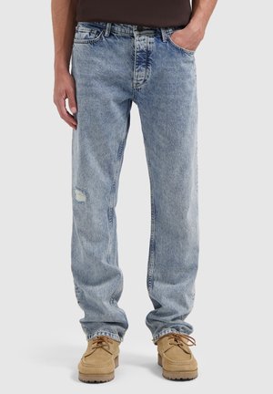 Man in lichtblauwe verwassen jeans met lichte slijtage op de linkerdij en tan suede veterschoenen tegen een witte achtergrond.