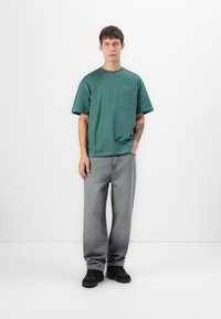 Green cotton T-shirt with a chest pocket, paired with loose gray denim jeans and black shoes, positioned against a plain white background.