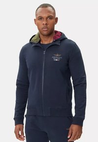 Felpa blu navy con cappuccio foderato in verde e bordeaux. Presenta polsini e orlo a coste, logo ricamato sul petto. Tessuto morbido al tatto.