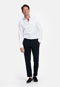 Witte button-up shirt met zwarte knopen, gecombineerd met zwarte op maat gemaakte broek en donkere schoenen. Man staat glimlachend tegen een effen achtergrond.