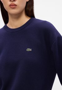 Námornícky pletený sveter s okrúhlym výstrihom a lemovanými manžetami, s malým zeleným krokodílím logom na ľavej hrudi.