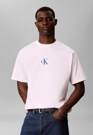 Ung mand iført hvid Calvin Klein T-shirt med blåt logo, mørkeblå jeans, sort bælte og sølvkæde-armbånd, stående foran grå baggrund.