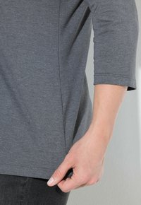De hand van een vrouw houdt de zoom van een grijze blouse met 3/4 mouwen tegen een lichte achtergrond, waarbij de textuur van de stof en de stiksels zichtbaar zijn.