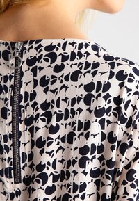 Kauluspaita, jossa on vaalea kangas kermanvärisenä ja abstrakteja tummanlaivastonsinisiä muotoja; sisältää näkyvän vetoketjun takana.