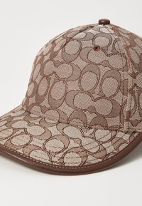 Gorra marrón con una tela de malla texturizada que presenta un patrón ovalado repetido. Acentos marrones brillantes en el ala y el botón superior.