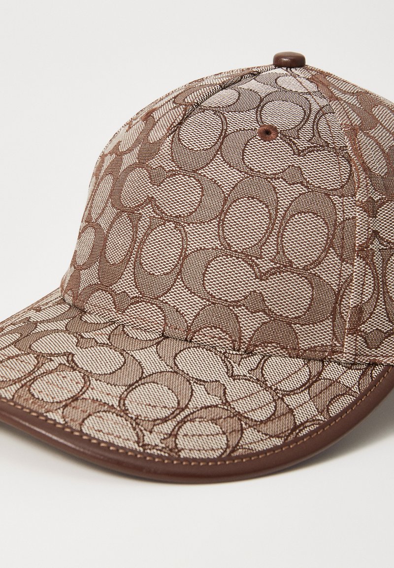 Gorra marrón con una tela de malla texturizada que presenta un patrón ovalado repetido. Acentos marrones brillantes en el ala y el botón superior.