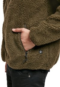 Veste en polaire de couleur vert olive foncé, avec un extérieur texturé, une poche zippée et un petit logo au bas.