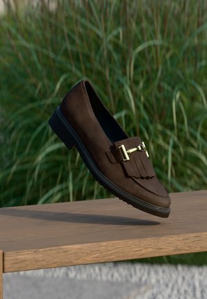 Dunkelbrauner Wildleder-Loafer mit goldener Schnalle und Fransen-Detail, der über einer Holzbank schwebt, grüner Gras-Hintergrund.