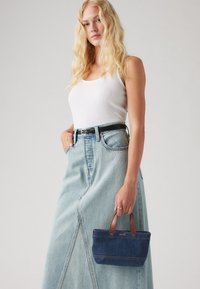 Τσάντα τζιν με καφέ χειρολαβές, με σκούρο μπλε ύφασμα και αντίθετη μπεζ ραφή, κρατημένη από ένα μοντέλο που φοράει ένα λευκό μπλουζάκι και φούστα ανοιχτού μπλε χρώματος.