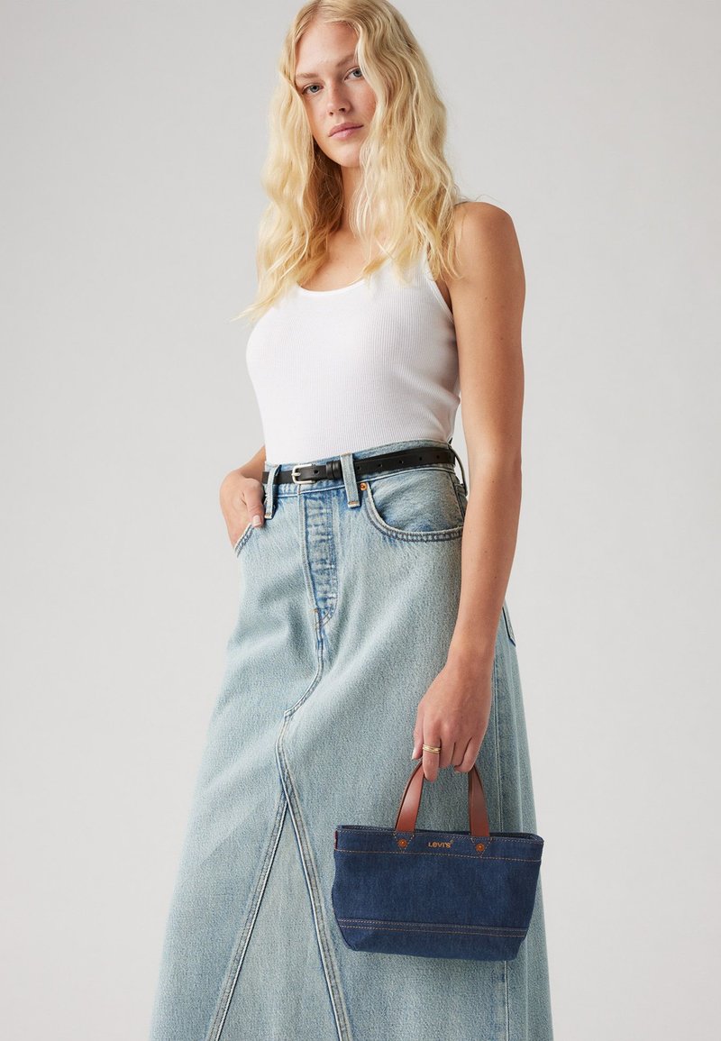 Τσάντα τζιν με καφέ χειρολαβές, με σκούρο μπλε ύφασμα και αντίθετη μπεζ ραφή, κρατημένη από ένα μοντέλο που φοράει ένα λευκό μπλουζάκι και φούστα ανοιχτού μπλε χρώματος.