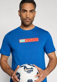 Μπλε βαμβακερό μπλουζάκι με κόκκινο και κίτρινο λογότυπο Nike BARÇA στο στήθος. Το άτομο κρατάει μια λευκή ποδοσφαιρική μπάλα με μπλε και μαύρες λεπτομέρειες.