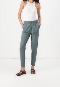 Helle grau-grüne Hose mit elastischem Bund, schmal zulaufendem Bein und seitlichen Taschen; kombiniert mit gemusterten flachen Schuhen und einem weißen Tanktop.